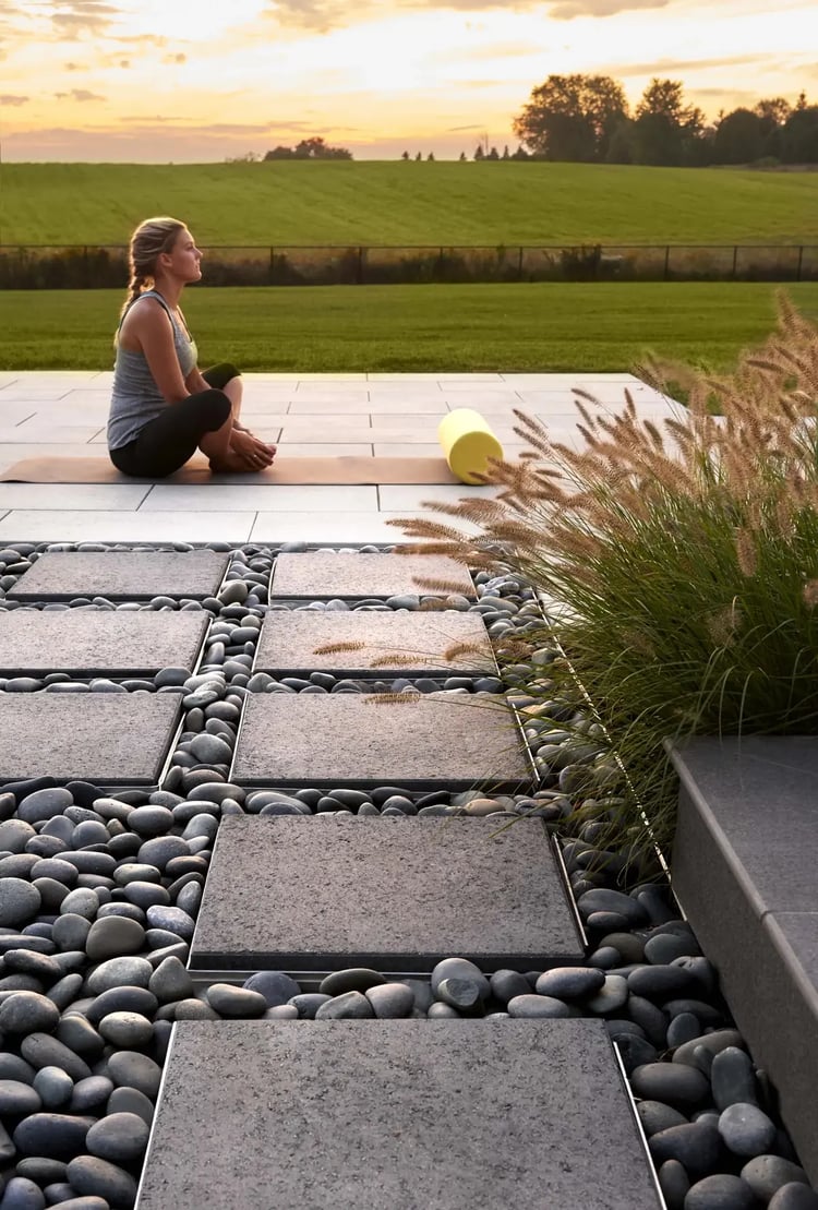 Patio moderno al aire libre con losas Para de Techo-Bloc e incrustaciones de rocas de río, con una sesión de yoga al atardecer con vistas abiertas a un campo verde y pastos ornamentales.
