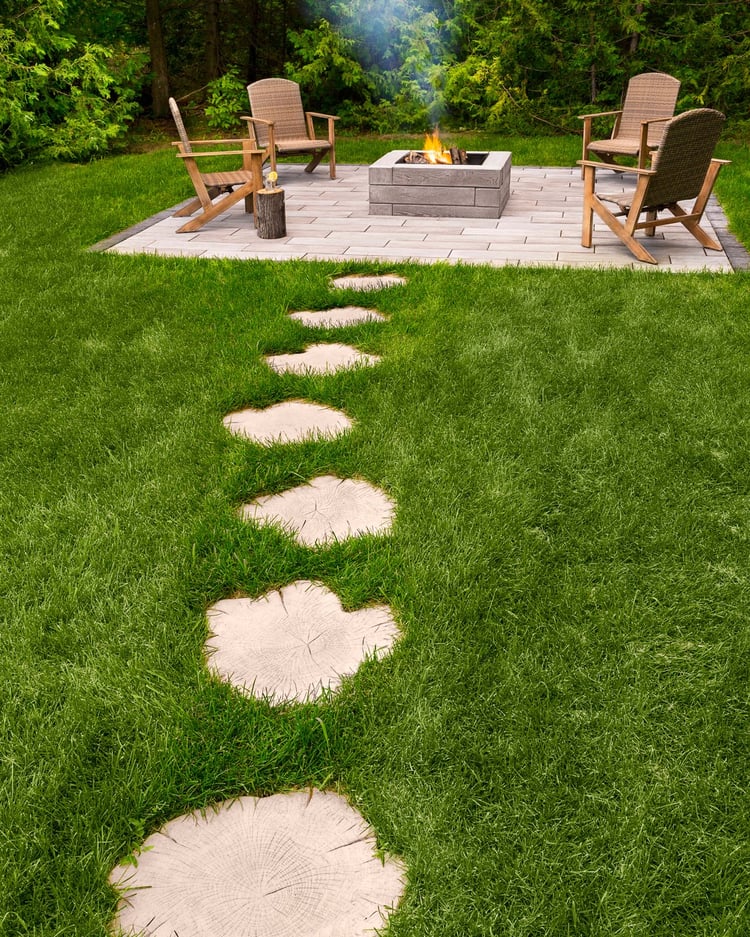 Adoquines Techo-Bloc Borealis con aspecto de madera natural que cruzan un exuberante césped hasta una acogedora área de fogata en el patio trasero con luces de cadena y sillas Adirondack.