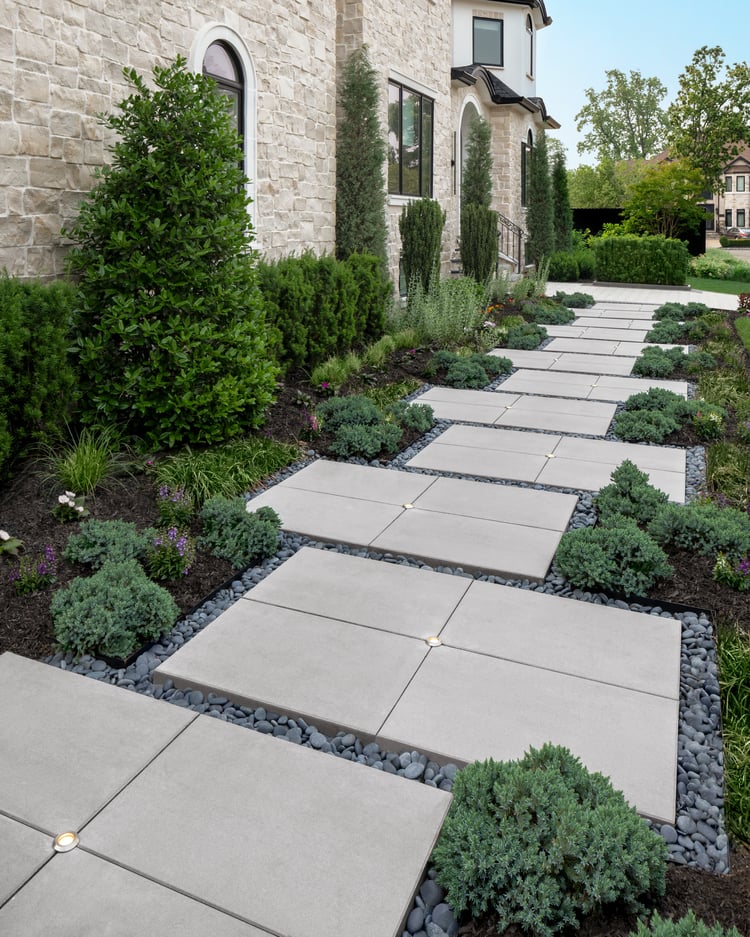 Pasarela delantera contemporánea con losas Techo-Bloc Para con iluminación integrada, rodeada de guijarros decorativos negros y exuberantes plantaciones de hoja perenne junto a una casa revestida de piedra.