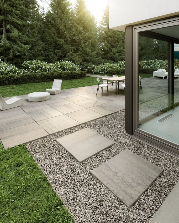 Patio moderno con losas de gran formato Techo-Bloc Industria 900 para el patio y senderos, rodeado de vegetación, detalles de grava y mobiliario exterior minimalista.