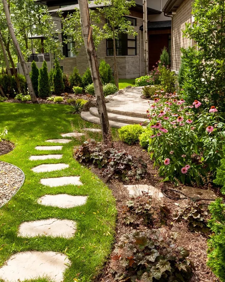 Escalones Techo-Bloc Maya establecidos en un camino de jardín curvado a través de un patio exuberante, lleno de flores, lo que lleva a un aterrizaje de adoquines y la entrada principal enmarcada por los árboles de abedul.
